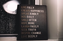 A motivational sign with various inspirational phrases such as 'LIVE FULLY', 'CREATE HAPPINESS', and 'LOVE DEEPLY' is placed next to a window with raindrops visible on the glass. The setting is dimly lit, creating a cozy and reflective atmosphere.
