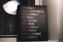 A motivational sign with various inspirational phrases such as 'LIVE FULLY', 'CREATE HAPPINESS', and 'LOVE DEEPLY' is placed next to a window with raindrops visible on the glass. The setting is dimly lit, creating a cozy and reflective atmosphere.