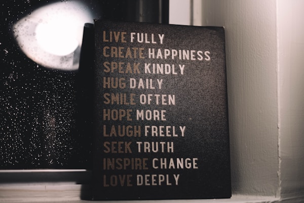 A motivational sign with various inspirational phrases such as 'LIVE FULLY', 'CREATE HAPPINESS', and 'LOVE DEEPLY' is placed next to a window with raindrops visible on the glass. The setting is dimly lit, creating a cozy and reflective atmosphere.