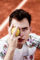 A person holding a yellow tennis ball up to one of their eyes while looking directly at the camera. They are wearing a white shirt with a floral pattern. The background is a clay tennis court with a visible white line.