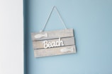 A decorative wooden sign hangs on a pastel blue wall. The sign consists of three horizontal wooden planks painted in a weathered, light gray color. White 3D text spelling 'Beach' is affixed to the center of the sign, flanked by two small, white fish decorations. The sign is suspended by a thin cord at the top.