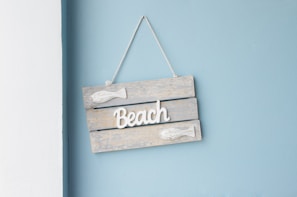 A decorative wooden sign hangs on a pastel blue wall. The sign consists of three horizontal wooden planks painted in a weathered, light gray color. White 3D text spelling 'Beach' is affixed to the center of the sign, flanked by two small, white fish decorations. The sign is suspended by a thin cord at the top.