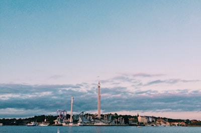 An amusement park is situated by the waterfront, with various rides and tall structures visible. The sky is mostly clear with some scattered clouds, and several boats can be seen on the water. Buildings line the shoreline, adding to the scenic landscape.