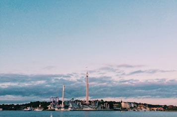An amusement park is situated by the waterfront, with various rides and tall structures visible. The sky is mostly clear with some scattered clouds, and several boats can be seen on the water. Buildings line the shoreline, adding to the scenic landscape.