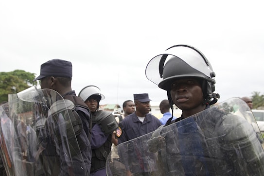 Several police officers wearing helmets and protective gear are standing together. Some are holding clear riot shields. They appear to be in a public outdoor setting, possibly during a demonstration or public event.