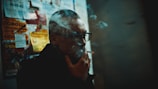 A person with glasses is smoking a cigarette, partially obscured by smoke. The individual is standing near a wall filled with various posters and advertisements. The setting appears dimly lit, with a moody, almost cinematic atmosphere.