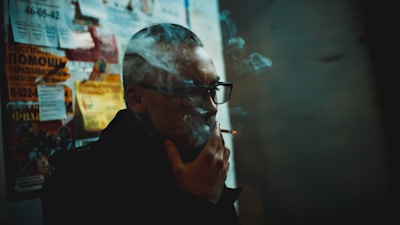 A person with glasses is smoking a cigarette, partially obscured by smoke. The individual is standing near a wall filled with various posters and advertisements. The setting appears dimly lit, with a moody, almost cinematic atmosphere.
