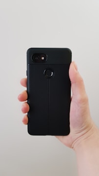 A hand is holding a smartphone that has a black leather-textured case. The phone features a camera and a fingerprint sensor on the back.