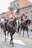 Several individuals are riding horses on a city street, dressed in traditional cowboy attire including wide-brimmed hats and decorative jackets. A Mexican flag is visible in the background, suggesting a cultural or festive event.