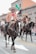 Several individuals are riding horses on a city street, dressed in traditional cowboy attire including wide-brimmed hats and decorative jackets. A Mexican flag is visible in the background, suggesting a cultural or festive event.