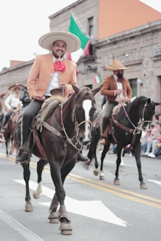 Several individuals are riding horses on a city street, dressed in traditional cowboy attire including wide-brimmed hats and decorative jackets. A Mexican flag is visible in the background, suggesting a cultural or festive event.
