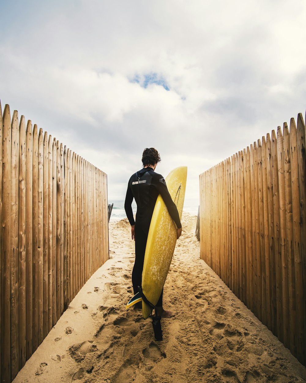 person standing between privacy fences while holding yellow surfboard during daytime