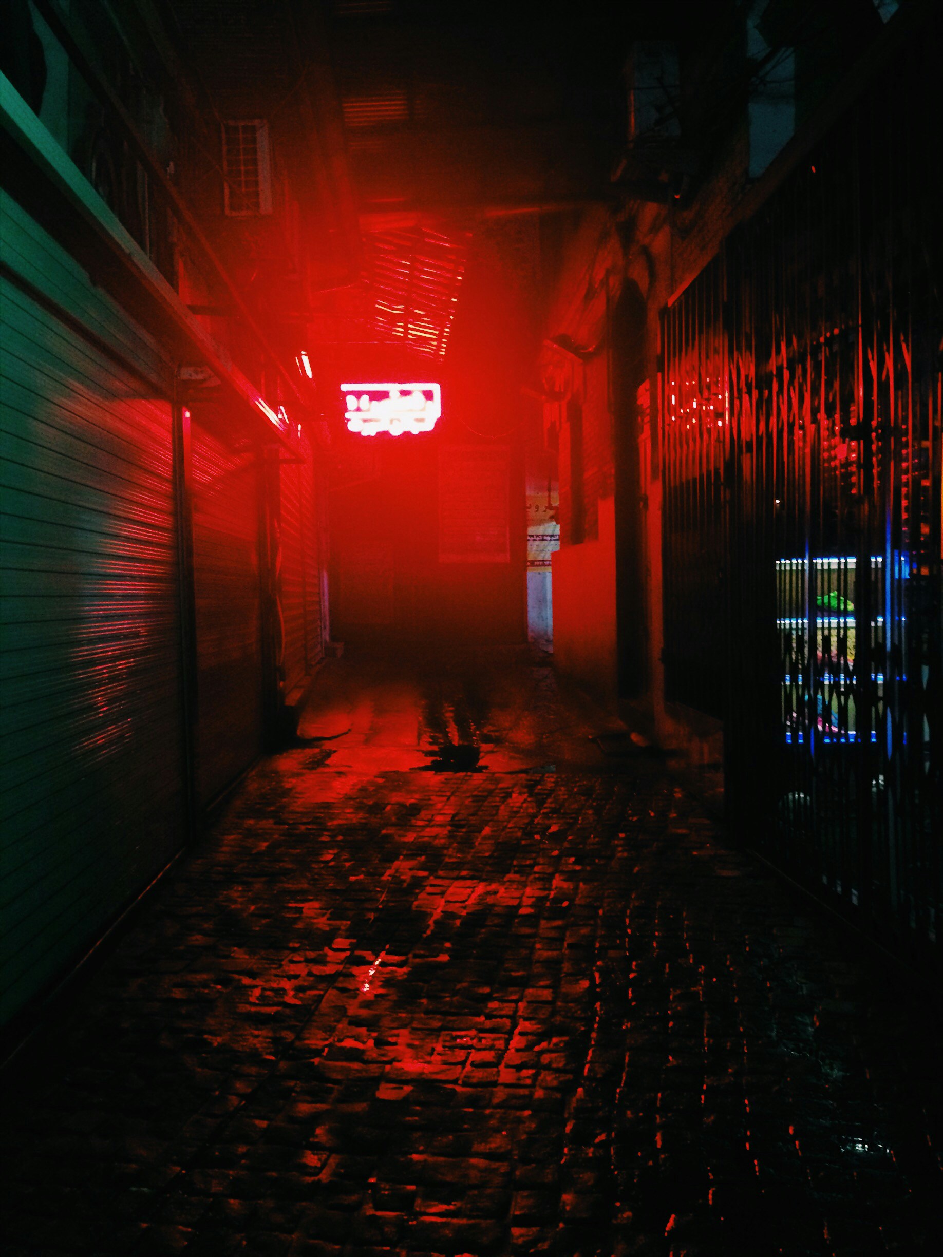 Dark urban alleyway at night with red neon signs reflecting on wet pavement and shadowy figures