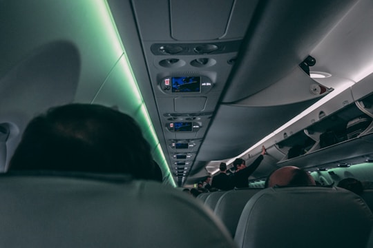 A view of an airplane cabin with overhead compartments and seats visible. Passengers are seated, and some individuals are standing or moving in the aisle. The lighting is dim with a soft green hue, contributing to a calm ambiance.