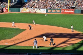 A baseball game in progress with players positioned on the field. The field is well-maintained with a crowd of spectators in the stands. Advertisement banners line the outfield fence, adding vibrant colors to the backdrop.