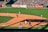 A baseball game in progress with players positioned on the field. The field is well-maintained with a crowd of spectators in the stands. Advertisement banners line the outfield fence, adding vibrant colors to the backdrop.