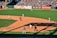A baseball game in progress with players positioned on the field. The field is well-maintained with a crowd of spectators in the stands. Advertisement banners line the outfield fence, adding vibrant colors to the backdrop.