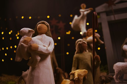 A wooden nativity scene with carved figures, including a person holding a lamb and others standing nearby. The background features soft, warm lights that create a cozy and serene atmosphere.