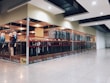 A modern clothing store with glass walls displaying a variety of garments on racks. There are mannequins positioned near the entrance dressed in different outfits. The store has bright lighting and a spacious tiled floor.