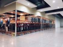 A modern clothing store with glass walls displaying a variety of garments on racks. There are mannequins positioned near the entrance dressed in different outfits. The store has bright lighting and a spacious tiled floor.