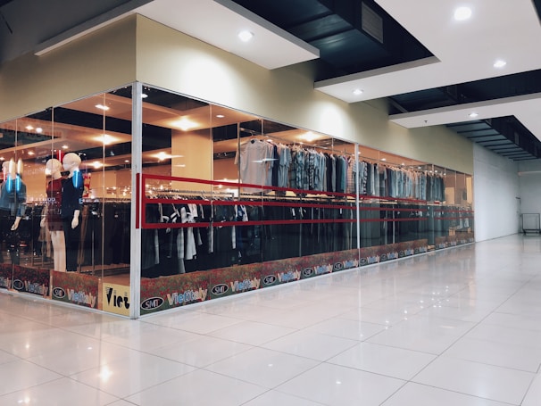 A modern clothing store with glass walls displaying a variety of garments on racks. There are mannequins positioned near the entrance dressed in different outfits. The store has bright lighting and a spacious tiled floor.