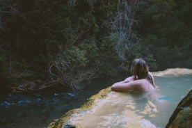 A person relaxes in a natural hot spring, gazing towards the lush, dense foliage of a forest. The water is calm and light-colored, contrasting against the dark green of the surrounding trees. The atmosphere is tranquil and secluded.