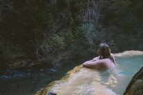 A person relaxes in a natural hot spring, gazing towards the lush, dense foliage of a forest. The water is calm and light-colored, contrasting against the dark green of the surrounding trees. The atmosphere is tranquil and secluded.