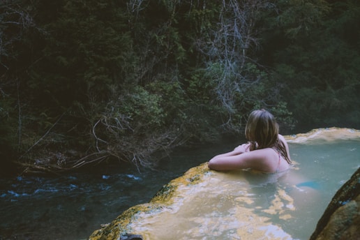 A person relaxes in a natural hot spring, gazing towards the lush, dense foliage of a forest. The water is calm and light-colored, contrasting against the dark green of the surrounding trees. The atmosphere is tranquil and secluded.