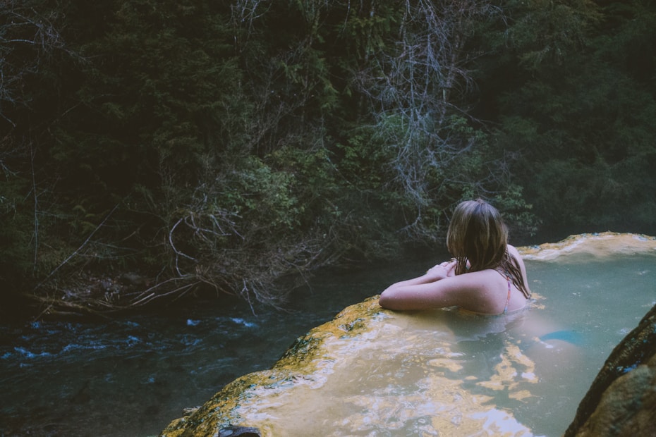 A person relaxes in a natural hot spring, gazing towards the lush, dense foliage of a forest. The water is calm and light-colored, contrasting against the dark green of the surrounding trees. The atmosphere is tranquil and secluded.