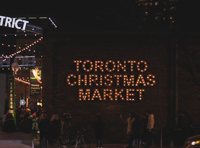 A nighttime scene depicts a bustling crowd gathered near a brick wall adorned with the illuminated words 'Toronto Christmas Market.' String lights hang overhead, adding warmth to the lively atmosphere. A variety of people, some standing and others walking, contribute to the festive ambiance.