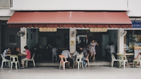 A small, casual eatery with a red awning and simple outdoor seating. Several people sit at plastic tables and chairs, some engaged in conversation or dining. A woman in a floral dress stands near the entrance. The ambience suggests a laid-back, everyday local dining experience.