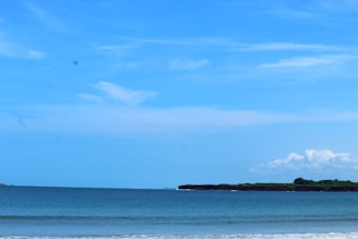 A serene seascape featuring a calm ocean with gentle waves meeting the sandy shoreline. The horizon is lined with a strip of lush green land, likely an island or a distant shoreline. Above, the sky is a clear vibrant blue with a few scattered fluffy white clouds.