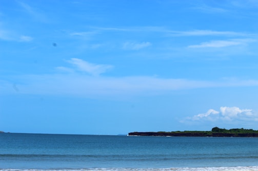 A serene seascape featuring a calm ocean with gentle waves meeting the sandy shoreline. The horizon is lined with a strip of lush green land, likely an island or a distant shoreline. Above, the sky is a clear vibrant blue with a few scattered fluffy white clouds.