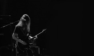 A musician with long hair is playing an electric guitar and standing in front of a microphone. The scene is dimly lit, creating a moody atmosphere with a focus on the performer.