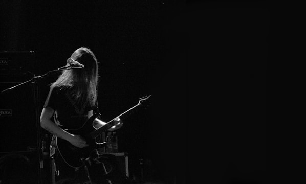 A musician with long hair is playing an electric guitar and standing in front of a microphone. The scene is dimly lit, creating a moody atmosphere with a focus on the performer.