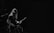 A musician with long hair is playing an electric guitar and standing in front of a microphone. The scene is dimly lit, creating a moody atmosphere with a focus on the performer.