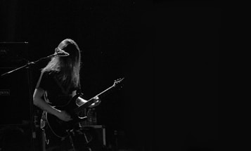 A musician with long hair is playing an electric guitar and standing in front of a microphone. The scene is dimly lit, creating a moody atmosphere with a focus on the performer.