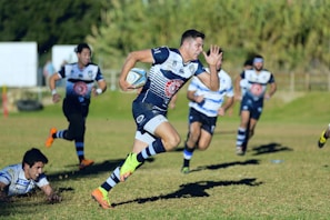A rugby player is sprinting across a grassy field with a rugby ball in hand. He is wearing a navy blue and white striped uniform, and one of his feet is visibly off the ground, indicating full motion. In the background, several other players can be seen, some wearing similar uniforms, while one player is on the ground reaching out as if attempting to tackle. The scene is set on a sunny day with green foliage in the background.