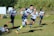 A rugby player is sprinting across a grassy field with a rugby ball in hand. He is wearing a navy blue and white striped uniform, and one of his feet is visibly off the ground, indicating full motion. In the background, several other players can be seen, some wearing similar uniforms, while one player is on the ground reaching out as if attempting to tackle. The scene is set on a sunny day with green foliage in the background.