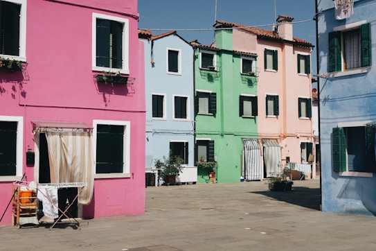 Colorful houses in a row, each with a distinct hue including pink, green, blue, and peach. The buildings are two to three stories high with dark green shutters and a few plants on window sills. The street in front is a quiet, sunlit square with some outdoor furniture and laundry hanging to dry.