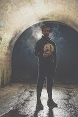 A person stands in a dimly lit tunnel holding a soccer ball. Smoke or fog swirls around them, creating a mysterious atmosphere. The tunnel walls are decorated with graffiti, and the ground appears wet, reflecting some light.