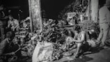 Several individuals are working amidst a cluttered pile of automotive parts and metal scraps, likely in a workshop or junkyard. The environment appears rustic and industrious, with various car engine components scattered around. The lighting creates a high-contrast, gritty effect.