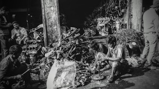 Several individuals are working amidst a cluttered pile of automotive parts and metal scraps, likely in a workshop or junkyard. The environment appears rustic and industrious, with various car engine components scattered around. The lighting creates a high-contrast, gritty effect.