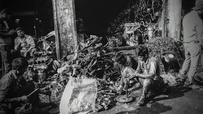 Several individuals are working amidst a cluttered pile of automotive parts and metal scraps, likely in a workshop or junkyard. The environment appears rustic and industrious, with various car engine components scattered around. The lighting creates a high-contrast, gritty effect.