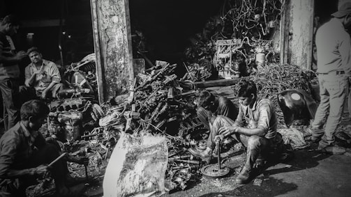 Several individuals are working amidst a cluttered pile of automotive parts and metal scraps, likely in a workshop or junkyard. The environment appears rustic and industrious, with various car engine components scattered around. The lighting creates a high-contrast, gritty effect.
