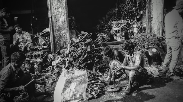 Several individuals are working amidst a cluttered pile of automotive parts and metal scraps, likely in a workshop or junkyard. The environment appears rustic and industrious, with various car engine components scattered around. The lighting creates a high-contrast, gritty effect.