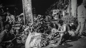 Several individuals are working amidst a cluttered pile of automotive parts and metal scraps, likely in a workshop or junkyard. The environment appears rustic and industrious, with various car engine components scattered around. The lighting creates a high-contrast, gritty effect.