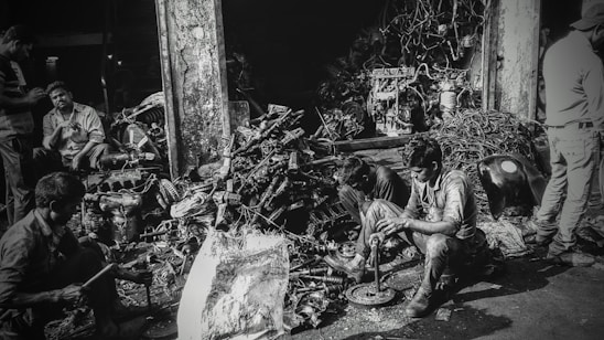 Several individuals are working amidst a cluttered pile of automotive parts and metal scraps, likely in a workshop or junkyard. The environment appears rustic and industrious, with various car engine components scattered around. The lighting creates a high-contrast, gritty effect.