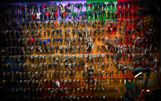 An array of various keys is neatly organized and displayed on a multicolored wall. The keys differ in shape and size, some with plastic grips while others are entirely metal. The background features a gradient of colors, ranging from red and green at the top to darker shades at the bottom. A lamp positioned in the lower right corner adds a warm glow to the scene, highlighting the texture and variety of the keys.