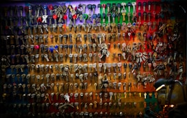 An array of various keys is neatly organized and displayed on a multicolored wall. The keys differ in shape and size, some with plastic grips while others are entirely metal. The background features a gradient of colors, ranging from red and green at the top to darker shades at the bottom. A lamp positioned in the lower right corner adds a warm glow to the scene, highlighting the texture and variety of the keys.